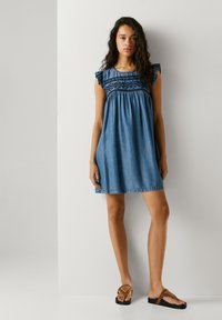 Ženska z dolgimi kodrastimi lasmi stoji obrnjena naprej in nosi modro brezrokavno obleko do kolen z vezenjem ter rjave platformne sandale.