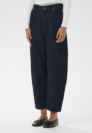 Person iført vidtbenede mørkeblå jeans, en hvid langærmet top og sorte balletsko, der står på en ensfarvet hvid baggrund.