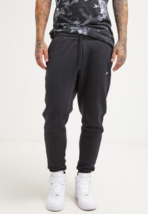 Mand iført sorte Nike joggingbukser og hvide høje sneakers, stående med synlige tatoverede arme mod en ensartet hvid baggrund.