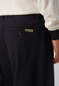 Pantalon noir avec une poche arrière boutonnée et une petite étiquette Marc O'Polo, porté avec une chemise blanche à manches longues côtelée.