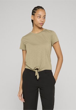 Giovane donna con i capelli intrecciati che indossa una camicia beige a maniche corte annodata e pantaloni neri, in piedi con una mano in tasca, guarda verso destra.