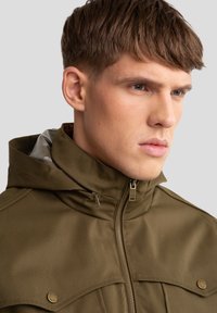 Olijfgroene jas met hoge kraag, lichte stof, ritssluiting en grote flapzakken, met zilveren hardware accenten.