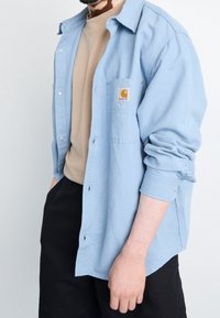 Mężczyzna w jasnoniebieskiej koszuli Carhartt z guzikiem, noszący beżowy sweter i czarne spodnie, z podwiniętymi rękawami koszuli.