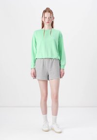 Felpa verde menta con maniche lunghe, abbinata a pantaloni corti a righe nere e bianche. Il modello indossa sneaker bianche e calzini bianchi a coste.