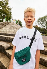 Groene schoudertas van stof met een ritszak, gedragen met een wit t-shirt met zwarte tekst. De achtergrond bestaat uit stenen trappen en groenvoorzieningen.