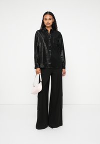 Camicia nera lucida a maniche lunghe con chiusura a bottoni, abbinata a pantaloni neri a gamba larga con tasche cargo. Porta una borsa rosa chiaro.