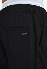 Fekete atlétikai rövidnadrág sima textúrával, hátsó zsebbel és oldalt ezüst "PEGADOR" logóval. Fehér csíkos derékpánt.