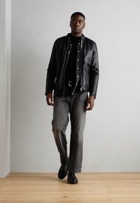 Zwarte leren jacket over een zwarte coltrui, gecombineerd met grijze flared jeans en zwarte enkellaarsjes. Model staat tegen een lichte muur.