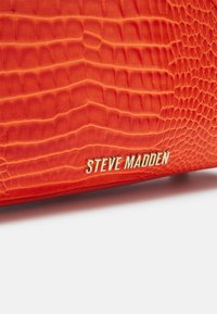 Sac à main orange en texture crocodile avec un logo « Steve Madden » en métal doré à l'avant, présentant une finition lisse.