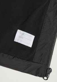 Interior de chaqueta polar negra con una etiqueta de tela blanca para nombre y teléfono. Presenta una cremallera negra y tela lisa y texturizada.