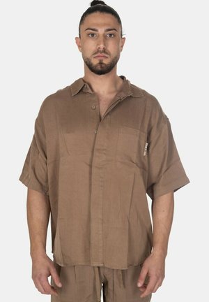 Uomo con capelli scuri raccolti indossa una camicia marrone a maniche corte, larga, con bottoni e pantaloni marroni abbinati, in piedi davanti a uno sfondo uniforme.