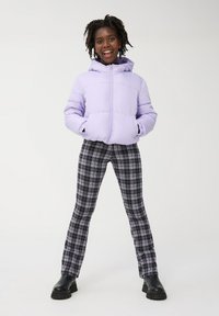Giacca a vento viola chiaro con cappuccio, pantaloni a quadri neri e viola, e stivali neri chunky, caratterizzati da una texture morbida e una vestibilità rilassata.