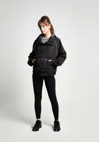 Mujer de pie con las manos en los bolsillos, vistiendo una chaqueta de viento negra, leggings negros y zapatillas deportivas negras, contra un fondo simple.