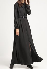 Robe maxi noire à manches longues, col avec nœud papillon, boutons sur le devant, taille ceinturée et fente latérale, en tissu lisse.