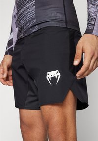 Zwarte sportshorts van rekbaar materiaal, met een witte cobra-logo aan de zijkant en een gespleten zoom voor flexibiliteit.