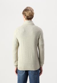 Pull en maille gris clair avec un col châle, un ourlet côtelé épais et un motif texturé, associé à un jean en denim bleu.