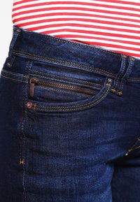 Jeans in denim scuro con una vestibilità attillata, dotati di una piccola tasca con zip, cuciture gialle a contrasto e leggere dettagli di texture.