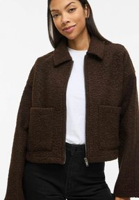 Brūna, teksturēta, īsā jaka ar apkakli, priekšpusē rāvējslēdzēju un divām uzšūtu kabatām. Valkāta pār baltu kreklu un melnām biksēm.