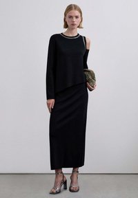 Top negro de manga larga con un escote adornado con detalles plateados, combinado con una falda negra. La modelo lleva sandalias de tacón alto plateadas con tiras.