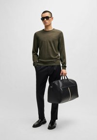 Schwarze, elegante Sporttasche mit einem weißen Reißverschluss, gehalten von einer Person, die ein olivgrünes langärmliges Shirt und schwarze, maßgeschneiderte Hosen trägt.