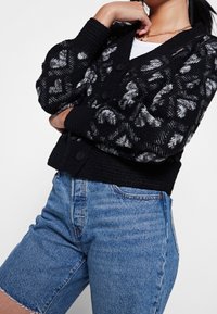 Nainen, joka on pukeutunut musta-valkoiseen kuvioituun neulekarvatuulettimeen valkoisen paidan ja korkeavyötäröisten sinisten farkkushortsien päälle, seisoo käsivarsia ristissä.