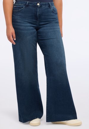 Jeans oscuros de pierna ancha combinados con zapatillas blancas, mostrando una vista parcial de la persona desde la cintura hasta los pies sobre un fondo liso.