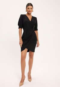 Czarna sukienka z opadającymi rękawami, z głębokim dekoltem w kształcie litery V i złotym guzikiem jako akcentem. Gładki materiał, dopasowana sylwetka i długość do kolan.
