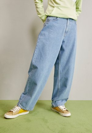 Personne portant un jean bleu clair à jambes larges, des baskets jaunes et beiges et un sweat-shirt vert clair debout sur un sol vert.