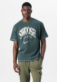 T-shirt in cotone verde scuro con una grande grafica bianca contenente il testo "SWOOSH" e il logo Nike, rifinito con cuciture a contrasto e una vestibilità rilassata.