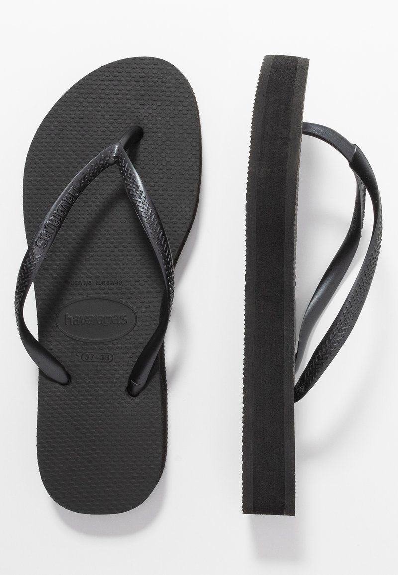 Mustat kumiset flip-flopit, joissa on teksturoitu pohja, V-muotoiset hihnat ja litteä pohja. Pohjassa on lievää kohotusta ja sileä reuna.