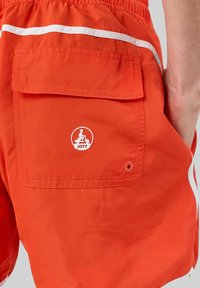 Pantaloni da bagno rossi con strisce laterali bianche, tasca posteriore con pattina e un logo circolare che raffigura una figura seduta, realizzati in materiale leggero.