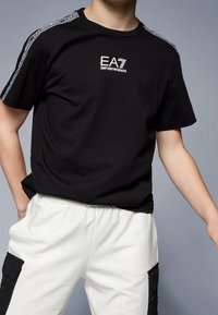 Černé bavlněné tričko s logem na hrudi a vzorovanými detaily na ramenou. Kombinováno s bílými tepláky s elastickým pasem.