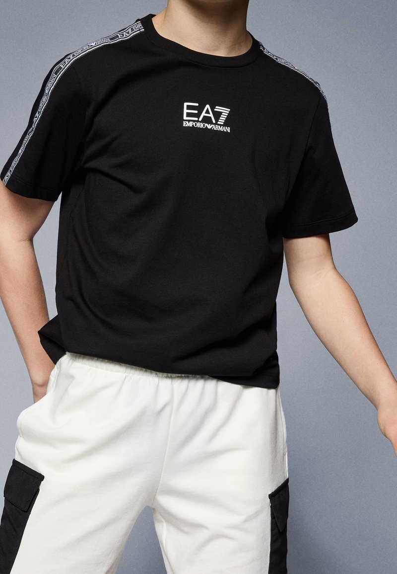 Černé bavlněné tričko s logem na hrudi a vzorovanými detaily na ramenou. Kombinováno s bílými tepláky s elastickým pasem.