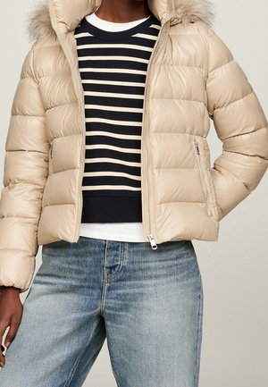 Personne portant une doudoune beige avec une capuche bordée de fourrure, un pull rayé noir et blanc, et un jean en denim bleu clair.