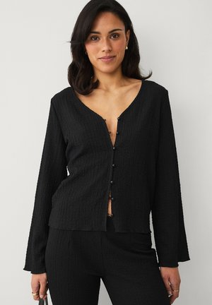 Kobieta o ciemnych włosach, ubrana w teksturowaną czarną koszulę na guziki i dopasowane spodnie, trzymająca małą czarną torebkę.