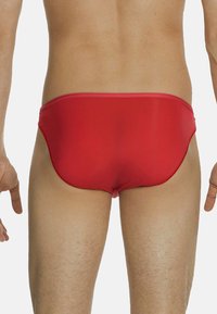 Schiena e gambe di un uomo che indossa slip da bagno rossi su sfondo bianco uniforme.