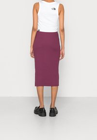 Vrouw in een witte mouwloze top met het The North Face-logo en een strakke paarse midi-jurk, staande met haar rug naar de camera op een effen achtergrond.