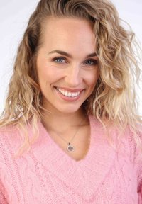 Roze kabelgebreide trui met een V-hals ontwerp, gecombineerd met een zilveren ketting met een ronde hanger. Het haar is golvend en blond.