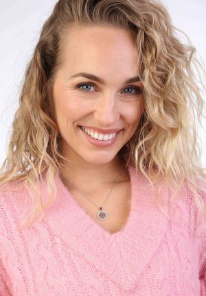 Roze kabelgebreide trui met een V-hals ontwerp, gecombineerd met een zilveren ketting met een ronde hanger. Het haar is golvend en blond.