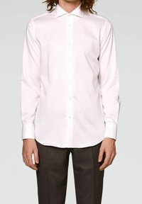 Chemise blanche à manches longues avec col boutonné, texture lisse, patte de boutonnage à l'avant et fermetures par boutons, portée avec un pantalon sombre sur mesure.