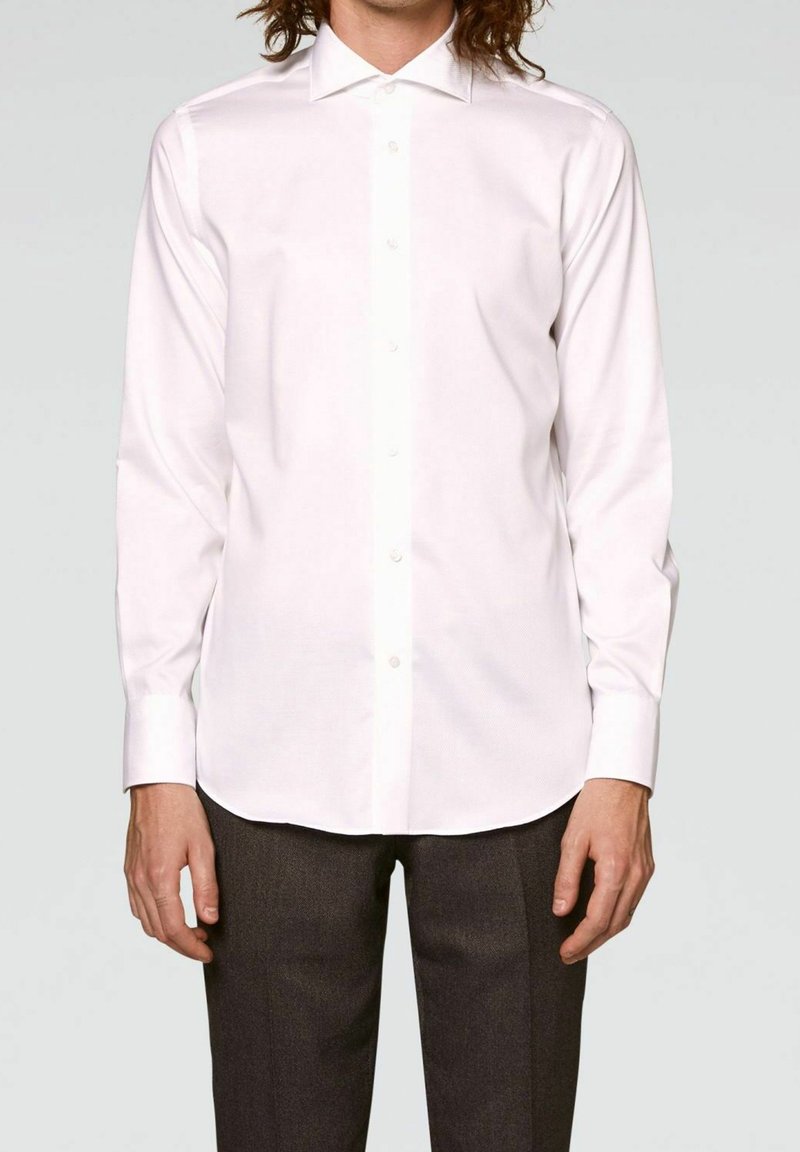 Chemise blanche à manches longues avec col boutonné, texture lisse, patte de boutonnage à l'avant et fermetures par boutons, portée avec un pantalon sombre sur mesure.