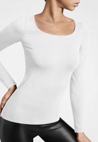 Langärmliges weißes Oberteil mit runder Ausschnittform und weicher, glatter Textur. Figurbetontes Design, kombiniert mit schwarzen Leggings. Minimalistischer Look.
