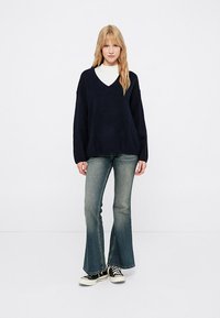 Tengerészkék kötött V nyakú pulóver laza szabással, választott koptatott farmernadrággal és fekete-fehér sportcipővel párosítva. Egyszerű és sík háttér.