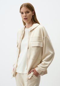 Beige zip-up hoodie met twee voorzakken, een trekkoordcapuchon en ribgebreide manchetten. Draagt over een wit t-shirt, onderdeel van een gecoördineerde outfit.