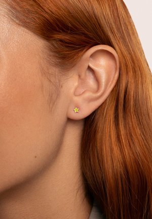 Pendiente de aro en forma de estrella dorada con un contorno verde, sujeto al lóbulo, exhibido en un modelo con el cabello rojo liso.