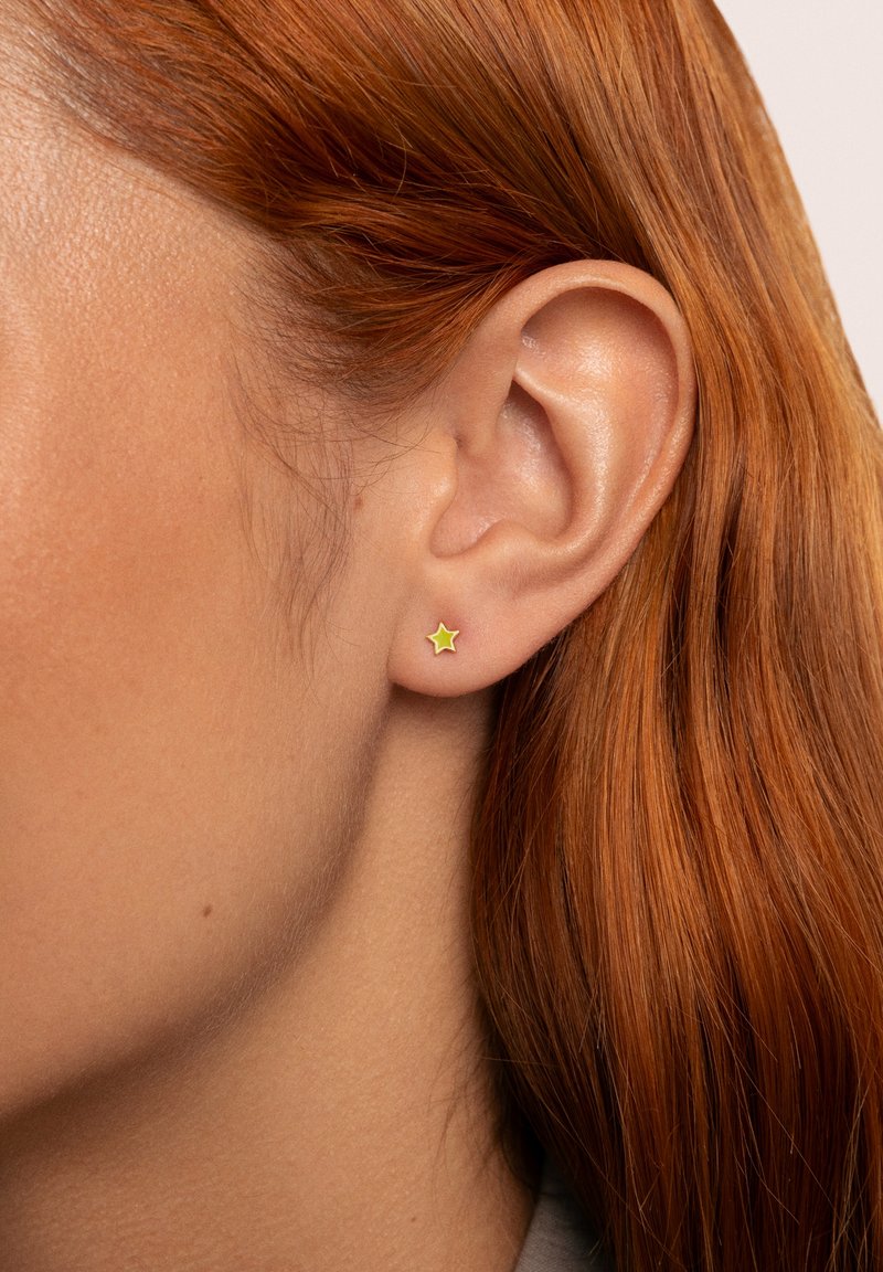 Pendiente de aro en forma de estrella dorada con un contorno verde, sujeto al lóbulo, exhibido en un modelo con el cabello rojo liso.