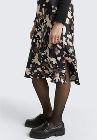 Zwarte jurk met een bloemenpatroon in crèmekleur, bruin en teal. Heeft een zijsplit en wordt gedragen met zwarte platform instappers en doorzichtige panty's.