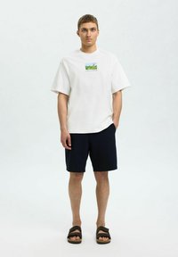 Biała bawełniana koszulka z krótkim rękawem i grafiką z przodu, połączona z granatowymi szortami i czarnymi sandałami. Prosty, casualowy strój.