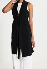 Schwarze ärmellose Weste mit Wickeldesign, taillierten Bund, Schalkragen und offener Vorderseite. Glattes Material, mittellang, minimalistischer Stil.