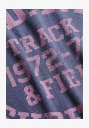 Granatowa koszulka z dużym różowym napisem "TRACK & FIELD 1972-73." Materiał wydaje się bawełniany, o miękkiej, teksturowanej powierzchni.